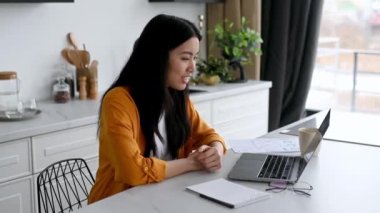 Dost canlısı, güzel Asyalı esmer kız, menajer, serbest çalışan, mutfakta oturuyor, dizüstü bilgisayar kullanıyor, müşteriyle ya da çalışanlarla görüntülü konuşuyor, projeyi tartışıyor, danışmanlık yapıyor, gülümsüyor.