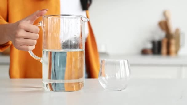 Una chica en ropa elegante casual, viene a la mesa en la cocina, vierte agua limpia de una jarra en un vaso, bebe una norma diaria de agua, cuida de su salud, lleva un estilo de vida saludable — Vídeos de Stock