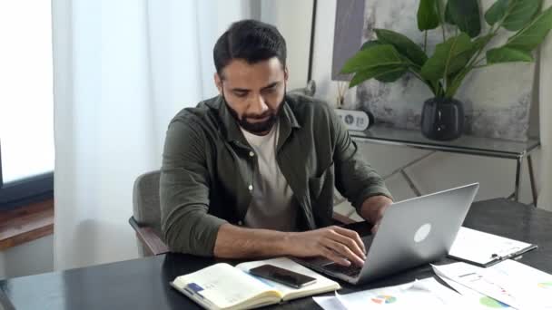 Indyjski lub arabski człowiek pozytywny, ubrany w stylowe ubrania, siedzący w swoim nowoczesnym biurze, rozmawiający przez telefon komórkowy z przyjacielem lub klientem podczas pracy na laptopie, prowadzący przyjazne negocjacje — Wideo stockowe