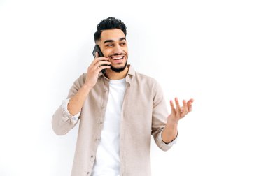 Neşeli, heyecanlı Hintli ya da Arap karizmatik adam hoş bir telefon konuşması yapıyor, mutlu yüz ifadesi, izole edilmiş beyaz arka planda duruyor, el kol hareketi yapıyor, gülümsüyor. Cep telefonuyla konuşma
