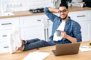 İş sırasında takılıyorum. Tarz sahibi Hintli ya da Arap bir adam, mutfakta bacakları masanın üzerinde oturuyor, elinde bir fincan kahve tutuyor, bilgisayarını kullanarak uzaktan çalışıyor.