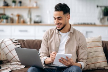 Pozitif zeki Hintli ya da Arap bir adam proje üzerinde çalışırken ya da çevrimiçi öğrenirken notlar alırken dizüstü bilgisayar kullanır, oturma odasında bir koltukta oturur, internette bilgi tarar, bir rapor hazırlar