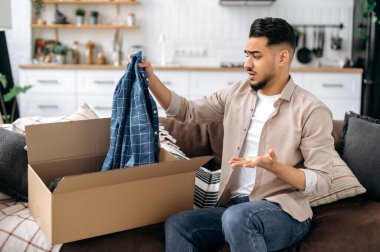 Üzgün Hintli ya da Arap adam büyük bir karton kutu açıyor, evdeki internet siparişini açıyor, uzun teslimat hizmetleri ve gelen mallar yüzünden hayal kırıklığına uğruyor,