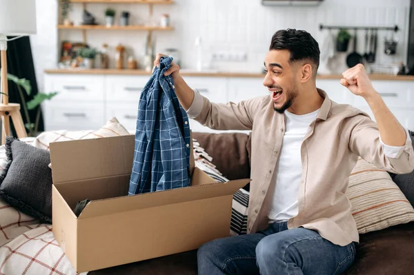 Mutlu Çekici Hintli ya da Arap adam büyük bir karton kutu açıyor, evde bir internet mağazası siparişi açıyor, hızlı teslimat hizmeti ve alınan, gülümseyen ve tatmin olan mallardan memnun.
