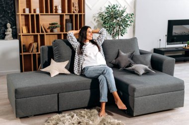 Uzun saçlı mutlu esmer kadın, ellerini başının arkasına koy, rahat bir koltukta modern bir oturma odasında otur, sakinliğin tadını çıkar ve hafta sonu evde tek başına otur, yan tarafa bak, gülümse.