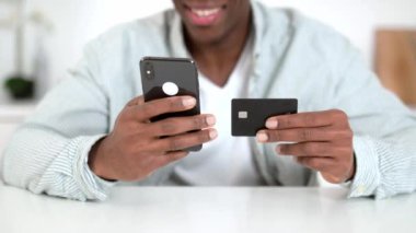 Cep telefonu ve kredi kartı erkeklerin elinde. İnternetten alışveriş yapan Afrikalı Amerikalı bir adam, akıllı telefon ve banka kartını kullanarak alışveriş yapıyor, veri girişi yapıyor.