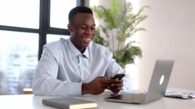 Olumlu çekici, genç, Afro-Amerikan iş adamı, menajer, modern ofiste iş yerinde oturuyor, akıllı telefon kullanıyor, iş arkadaşları veya müşterileriyle telefonda konuşuyor, arkadaşça gülümsüyor.