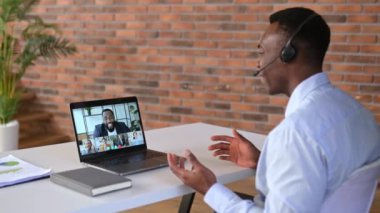 Sanal toplantı, iş arkadaşlarının beyin fırtınası, online sohbet. Afrikalı Amerikalı adam bilgisayar ekranına bakıyor, çok ırklı iş grubuyla görüntülü konuşma yapıyor, projeyi tartışıyor, selamlıyor.