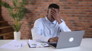 Sıkılmış Afrikalı Amerikalı bir adam, serbest çalışan ya da bilişim uzmanı, ofiste çalışıyor, bilgisayarla bir masada oturuyor, çok çalışıyor, yorgun, stresli bir durumda, dinlenmeye ihtiyacı var.