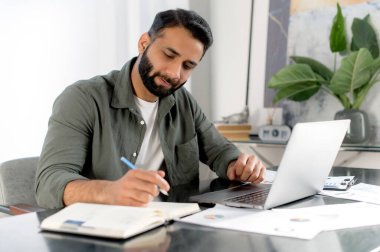 Arap ya da Hint uyruklu zeki, meşgul bir adam, başarılı şirket CEO 'su, finans müdürü, modern ofiste masasında oturuyor, belgelerle ve laptopla çalışıyor, analiz ediyor, notlar alıyor.