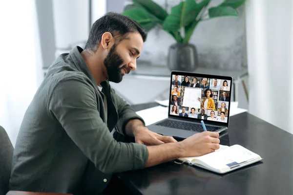 Side view of a successful smart guy listening to an online lecture, taking notes in a notebook, on a laptop screen, a teacher and a group of multiracial people. Online training, webinar