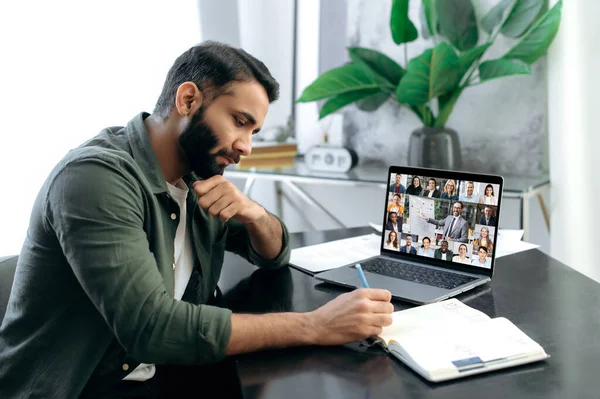 Financial educational course. Focused successful smart mixed race man listening to an online financial lecture, taking notes in notebook, on a laptop screen,a teacher and a group of multiracial people