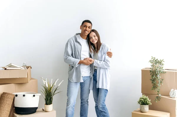 在他们的新公寓里 一对多种族兴奋的情侣站在白墙边 站在纸板箱之间 看着相机 开心地微笑着 — 图库照片