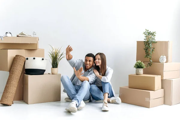 Moving to new apartment, buying home. Happy young married multiracial couple sitting on a floor nera white wall and between cardboard boxes with things, plans to repair and a project of new apartment