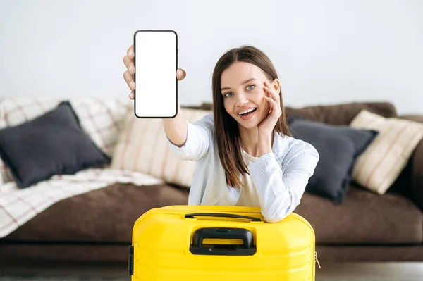Wireless technology. Joyful blue-eyed caucasian girl, sits in the living room near a yellow suitcase, shows a smartphone with a blank white mockup screen for advertise, looks at the camera, smiles