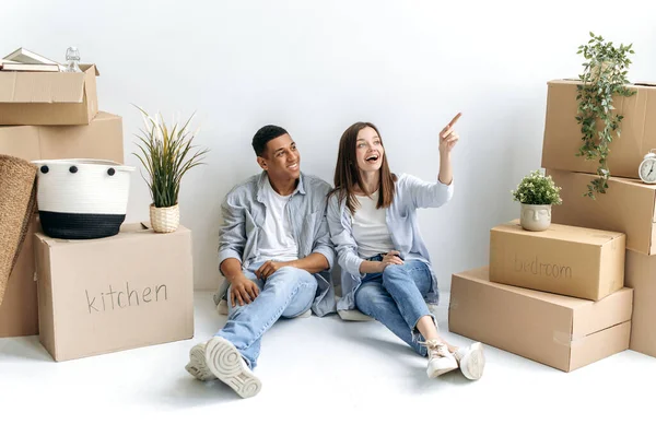 Happy young married multiracial couple sitting on a floor nera white wall and between cardboard boxes with things, plans to repair and a project of new apartment. Moving to new apartment, buying home