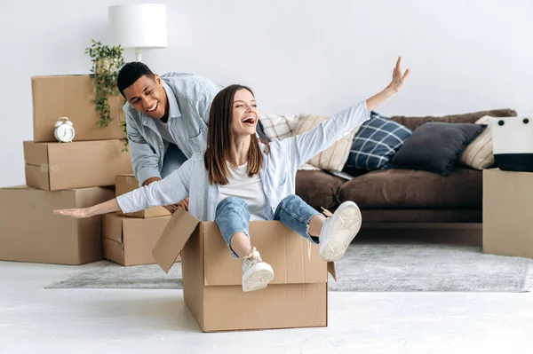 New home, relocation. Happy mixed race young family couple, bought their own house, they having fun on moving day, joyful wife sitting in cardboard box and husband push her, they laughing together
