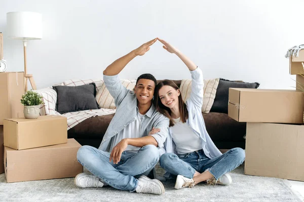 快乐快乐的多种族年轻夫妇相爱地坐在他们新家客厅的地板上 坐在纸板箱之间 手拿着一个假想的屋顶 看着相机 面带微笑 — 图库照片