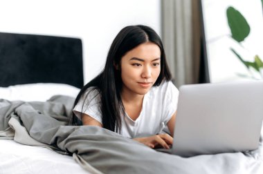 Kablosuz teknoloji. Pozitif Çinli esmer kız, beyaz tişörtlü, bir odada bir yatakta yatıyor, laptopunu kullanıyor, sosyal medyada arkadaşlarıyla sohbet ediyor, e-posta ve haberleri kontrol ediyor, gülümsüyor.