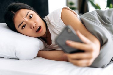 Sersemlemiş Çinli ya da Japon bir kız sabah yatakta yatarken, akıllı telefonuna şok olmuş gibi görünüyor, çalar saati fazla kaçırmış, duygusal yüz ifadesi, olumsuz duygu, beklenmedik haberler ya da mesaj.
