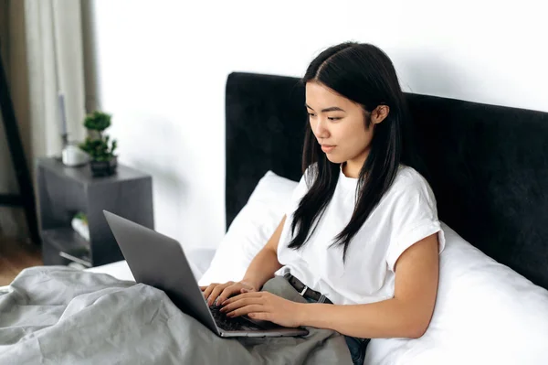 Lovely happy asian brunette girl dressed in a basic white t-shirt, is sitting in a bed in modern bedroom, uses her laptop, chatting with friends in social media, checking email, browsing news, smiling