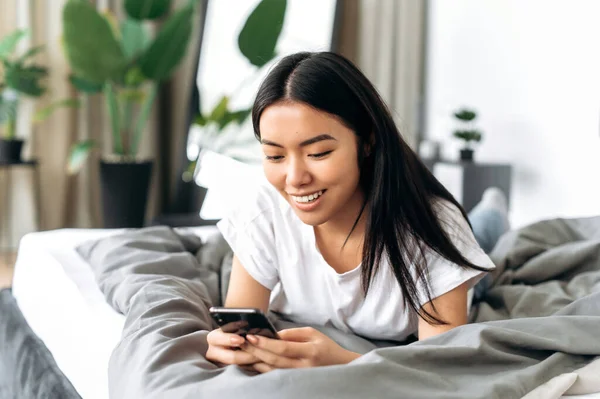 Pretty happy asian brunette girl dressed in a basic white t-shirt, is laying on a bed in a room, uses her cell phone, chatting with friends in social media, checking email, browsing news, smiling