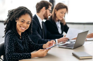 Afro-çekici Afro-Amerikan bir kadının resmi, tişörtlü, finans müdürü, ofiste iş arkadaşlarıyla bir masada oturuyor, kameraya bakıyor, dostça gülümsüyor.