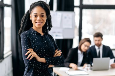Pozitif özgüvenli bir Afro-Amerikan iş kadını portresi, resmi gömlekli, kreatif direktör, iş lideri, modern bir ofiste kollarını kavuşturmuş, kameraya bakıyor, dostça gülümsüyor.