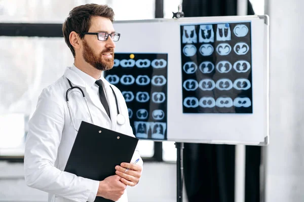 Proud pensive caucasian male general medicine doctor, surgeon, pulmonologist, in medical uniform, with glasses and stethoscope on shoulders, stand in the clinic, looks to the side, thinking