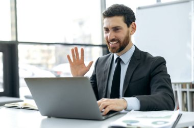 Sakallı, mali analist, şirket CEO 'su, modern bir ofiste dizüstü bilgisayarın önünde oturan iş arkadaşlarıyla video görüşmesi yapan, onları selamlayan, gülümseyen beyaz bir adam.