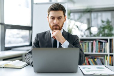 Odaklanmış zeki beyaz erkek, pazarlamacı, web geliştirici, finans müdürü, bir dizüstü bilgisayarın önünde, modern bir ofiste şık bir iş elbisesi içinde, bir dizüstü bilgisayarın içinde çalışıyor.