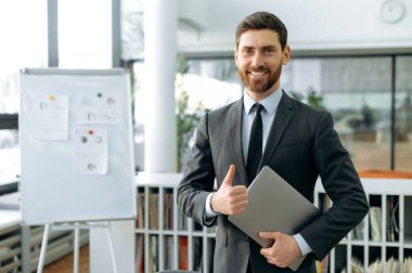 Zarif beyaz iş adamı, şirket CEO 'su, denetçi, modern bir ofiste, resmi bir iş elbisesi içinde elinde bir dizüstü bilgisayarla kameraya bakıyor, gülümsüyor, baş parmak işareti yapıyor.