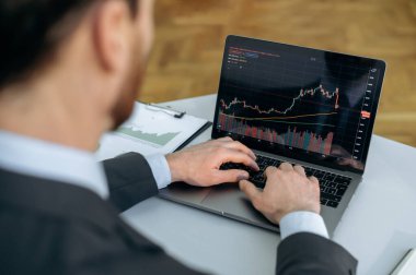 Borsa ticareti. Başarılı beyaz iş adamı, borsa simsarı, masasında oturuyor, dizüstü bilgisayar kullanıyor, tırnak içinde çalışıyor, riskleri ve olası kârları değerlendiriyor, bir strateji geliştiriyor.