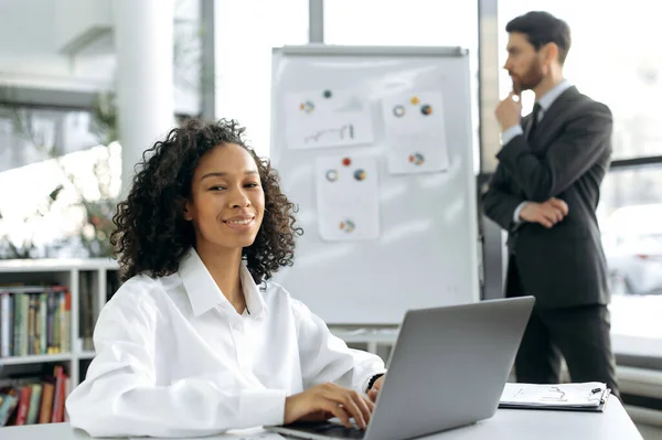 Smart positive dark-skinned businesswoman, web designer, sits at her desk in front of a laptop. In the background, a male colleague works with graphs, develops a strategy for modernizing the workflow