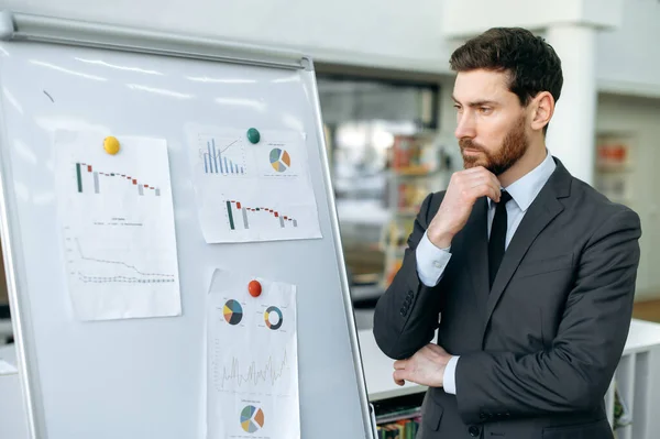 Focused elegant young adult caucasian man, company top manager, financial analyst, risk manager, stands near whiteboard with graphs in a modern office, concentrated looks at them, analyzes dynamics