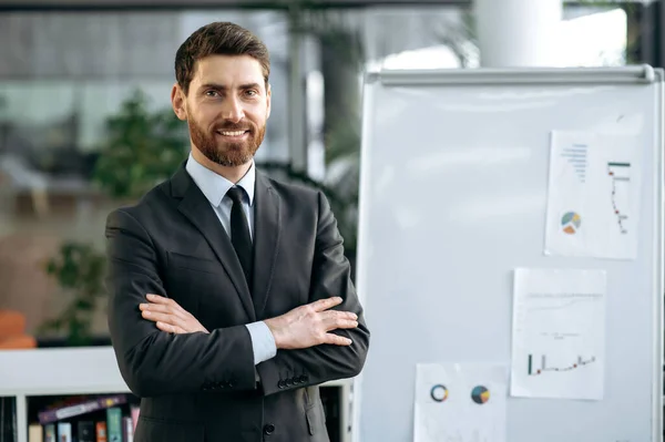 Respected intelligent Caucasian male entrepreneur, stock market broker, stands near whiteboard with charts in modern office, wearing business attire, arms crossed, looking at camera, smiling friendly