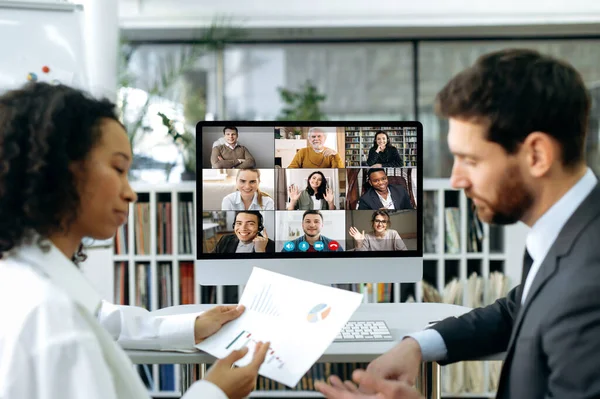 Company co-founders, a caucasian man and an african american woman, sit in a modern office in front of a computer screen, have an online video meeting with colleagues, demonstrate company profit