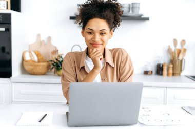Güzel, pozitif ırk kıvırcık bir kızın portresi. Günlük giysiler içinde, tasarımcı, serbest çalışan, dizüstü bilgisayarlı bir masada oturan, uzaktan çalışan, kameraya bakan, arkadaşça gülümseyen.