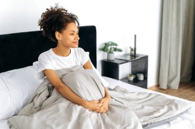 Yeni bir günün başlangıcı. Beyaz tişörtlü güzel Afro-Amerikan kıvırcık kız, iyi uyudu, rahat yatak odasında sabahları oturuyor, pozitif düşüncelerle yeni bir güne başlıyor ve gülümsüyor.