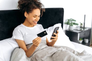 Afro-Afro-Amerikan kıvırcık kız, evde bir yatakta uzanıyor, akıllı telefon ve kredi kartıyla internetten mal alıyor, internetten sipariş veriyor. Çevrimiçi alışveriş, çevrimiçi dağıtım, teknoloji kavramı