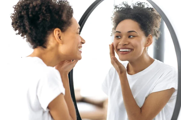 Positive pretty of African-American ethnicity girl with curly hair in a white t-shirt, stands in front of a mirror, examines her face, rejoices at the healthy state of her facial skin, smiling happily