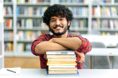 Pozitif zeki Hintli ya da Arap adam, karışık ırk erkek, üniversite öğrencisi, kitaplarla dolu bir masada oturuyor, kütüphanenin arka planında, kameraya bakıyor, arkadaşça gülümsüyor.