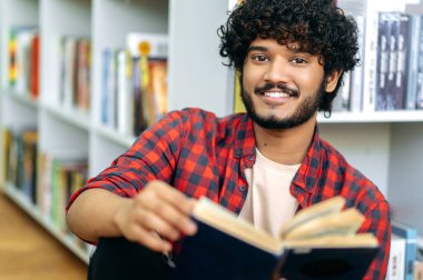 Yakışıklı, çekici, pozitif Hintli ya da Arap bir adam, kütüphanenin yanında oturuyor, elinde bir kitap tutuyor, okuyor, bilgi topluyor, sınava hazırlanıyor, kameraya bakıyor, arkadaşça gülümsüyor.