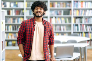 Yakışıklı, kendine güvenen kıvırcık ırklı adam, Hintli ya da Arap erkek, günlük giyim, öğrenci, serbest çalışan ya da öğretmen, kütüphanede arka planda duruyor, kameraya bakıyor, gülümsüyor.