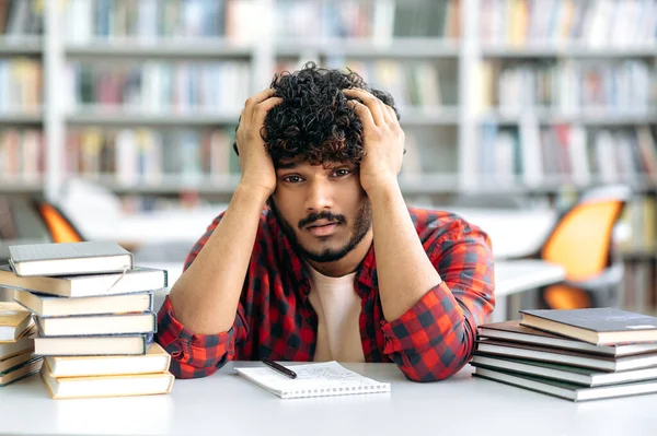 Tired exhausted indian or arabian guy, in casual clothes, a university student, or a high school student, sits at a desk in a library between books, holds his hands on his head, looks tired of reading