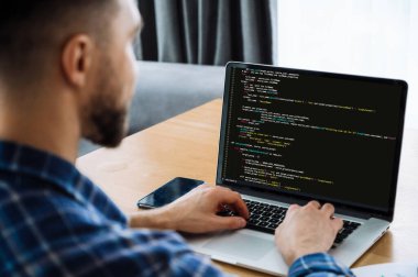 Kodlama teknolojileri geliştirmek, bilişim konsepti. Ekranda programlama kodu olan bir dizüstü bilgisayara erkek bilişim uzmanının omuz üstü görüntüsü. Yeni yazılım uygulaması üzerinde çalışan akıllı erkek programcı
