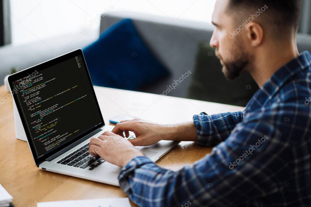Software concept. Side view of smart caucasian IT specialist, programmer, sits at home, uses laptop, concentrated working on the development of a website, application, writing codes and data code