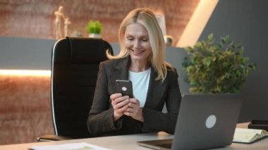 Joyful successful attractive influential mature caucasian businesswoman or top manager using cellphone while sitting at desk in office, texting client, friends or colleagues, got good news, messaging
