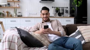 Moda kıyafetleri içindeki yakışıklı Hintli ya da Arap bir adam, modern bir oturma odasında rahat bir koltukta oturuyor, akıllı telefonunu tutuyor, mesaj atıyor, sosyal medyada geziniyor, internette sörf yapıyor, gülümsüyor.