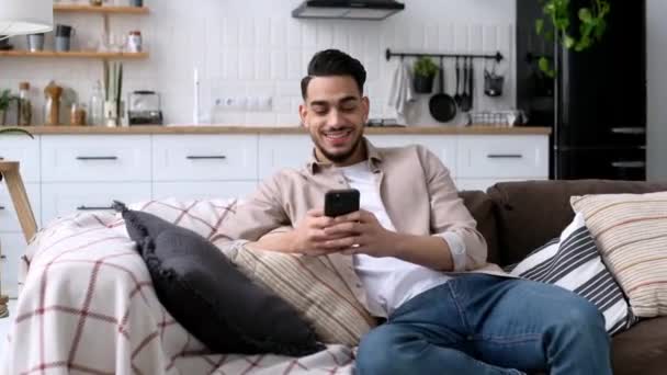 肯定的なハンサムなインド人やアラビア人の男流行の服で 快適なソファの上にモダンなリビングルームに座って 彼のスマートフォン テキストメッセージを保持し ソーシャルメディアを閲覧し ウェブサーフィン — ストック動画